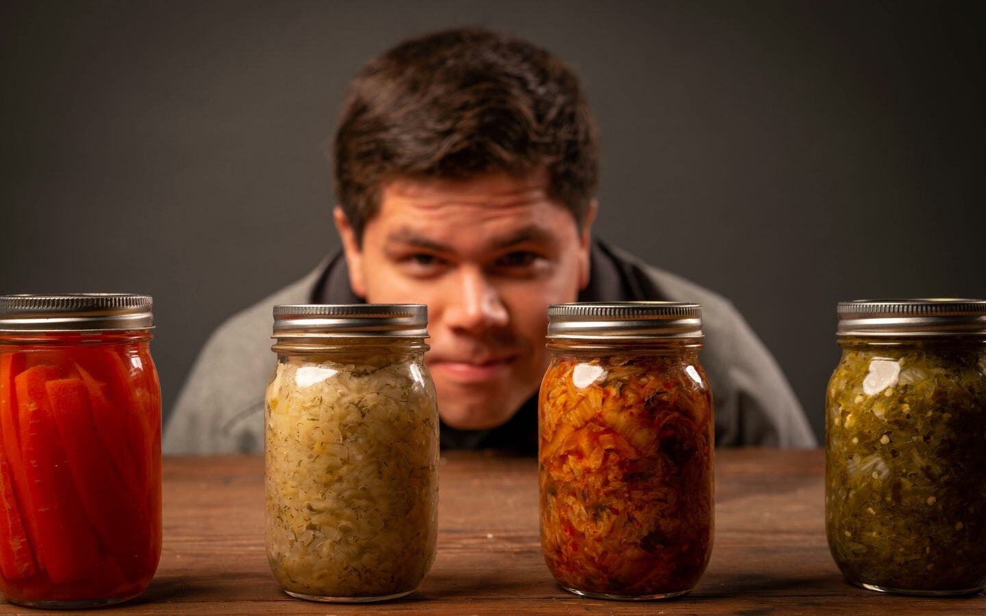 A man bending and looking at 4 jars of fermented foods sat on top of a wooden table