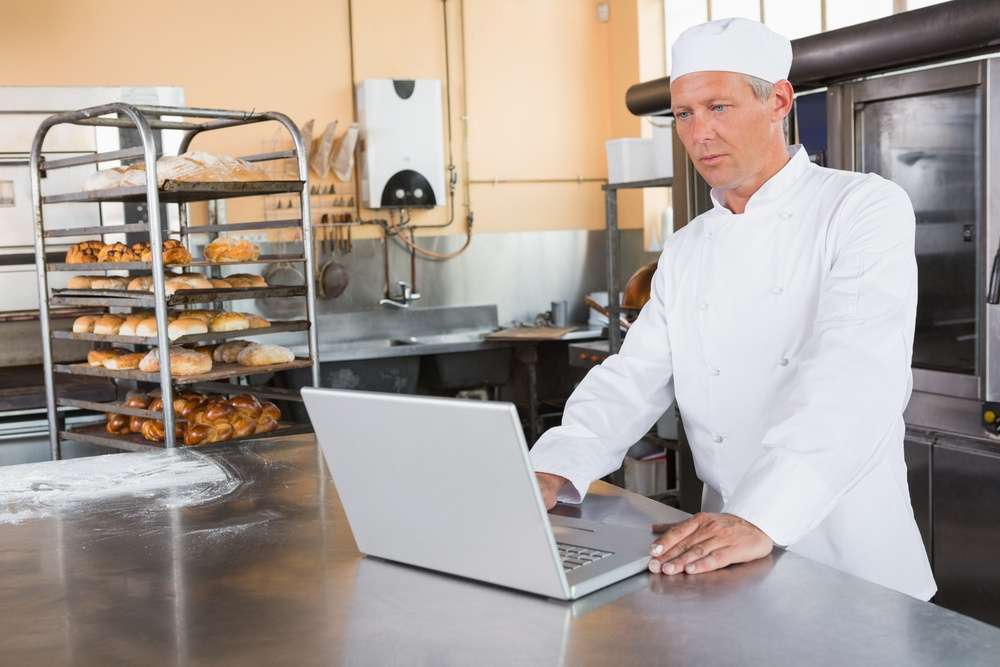 Focused baker using laptop on worktop in the kitchen of the bakery Focused baker using laptop on worktop in the kitchen of the bakery