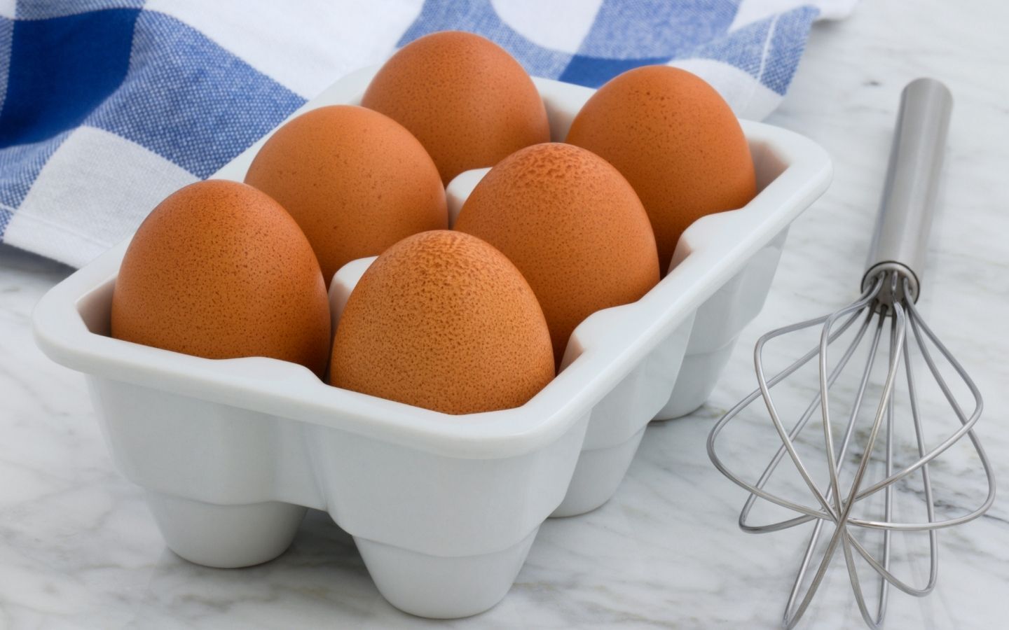 6 eggs sitting in an egg tray with a whisk sat beside it and a blue and white chequered tea towel behind