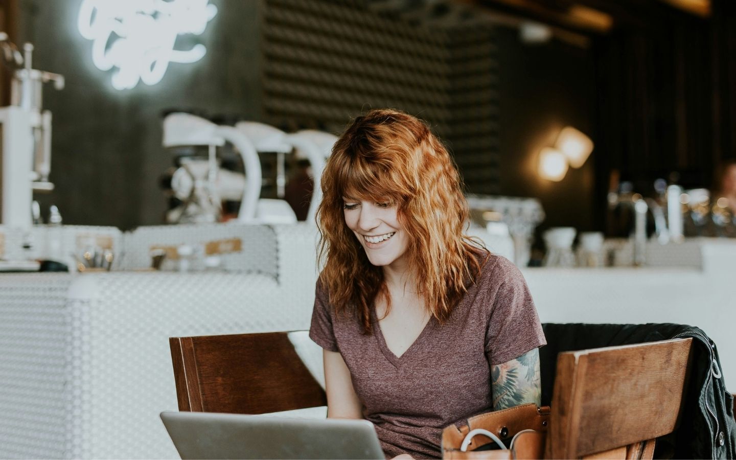 A smiling red-headed woman with tattoos sat in a cafe with her laptop