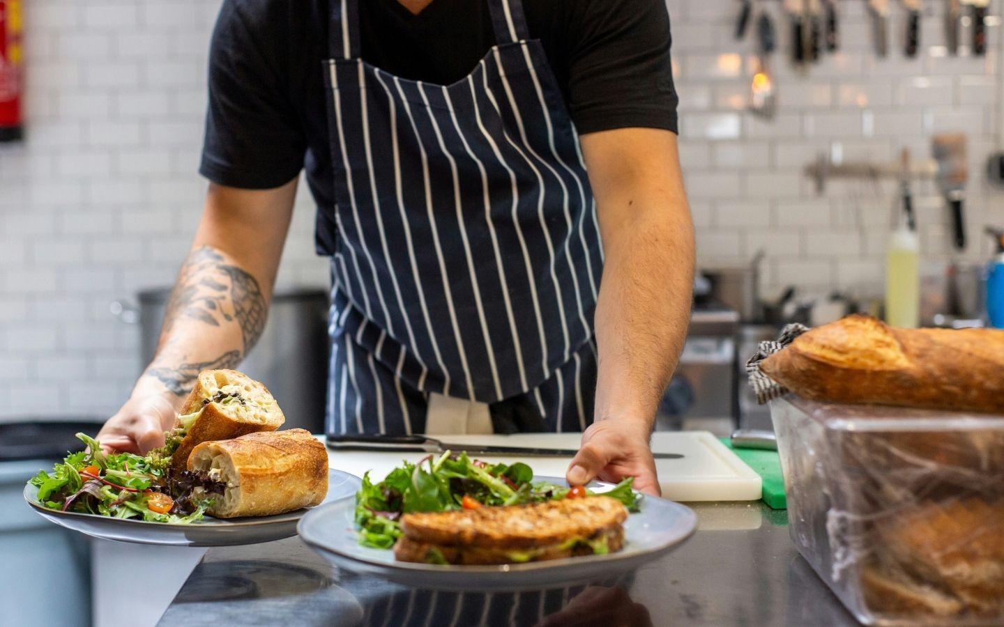 A chef in a stripey apron serving up two plates with toasting sandwiches and a side salad