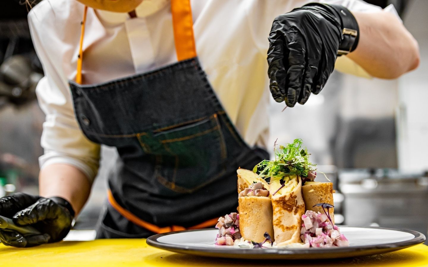 A person wearing an apron and gloves sprinking on micro greens on a plate of wraps and salad