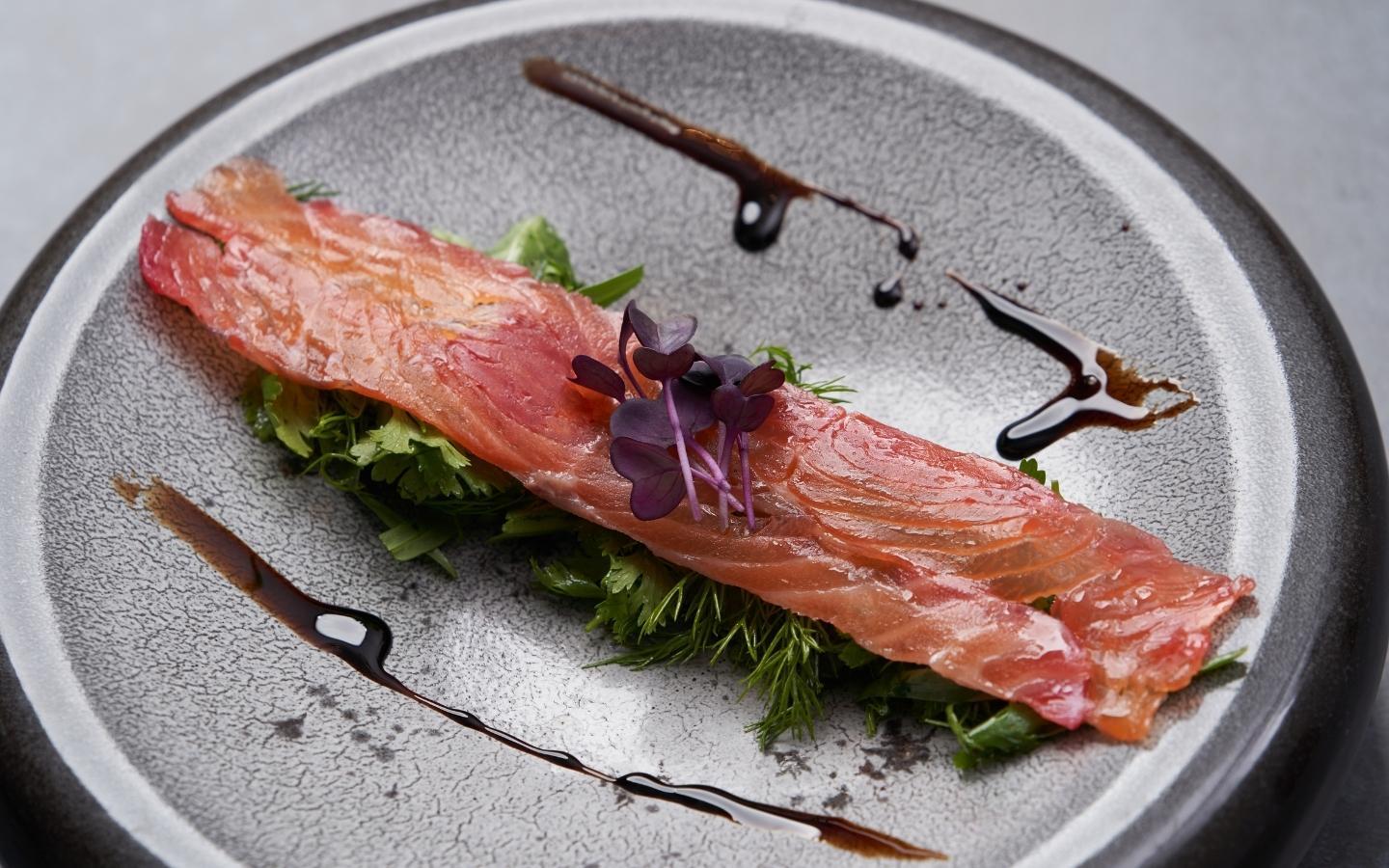 A grey plate with a pieve of smoked salmon on it garnished with herbs and a dressing